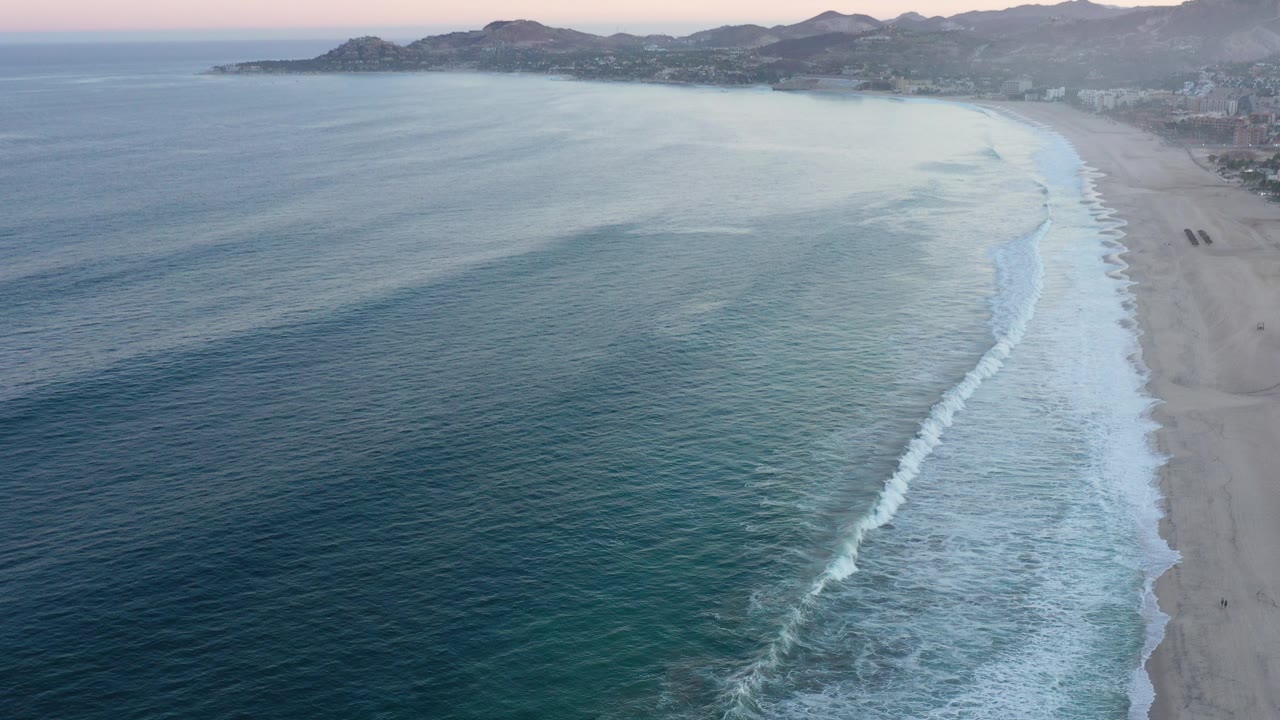 Aerial dusk view of San Jose del Cabo shoreline with blue-green water on the left, a pale beach on the right, distant mountains, and no visible people.