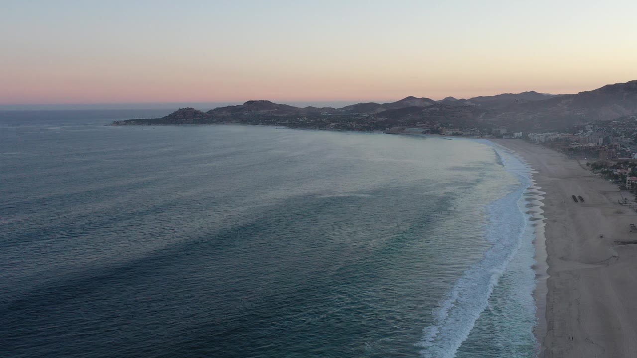 Aerial view of ocean waves beside a long beach at sunset.