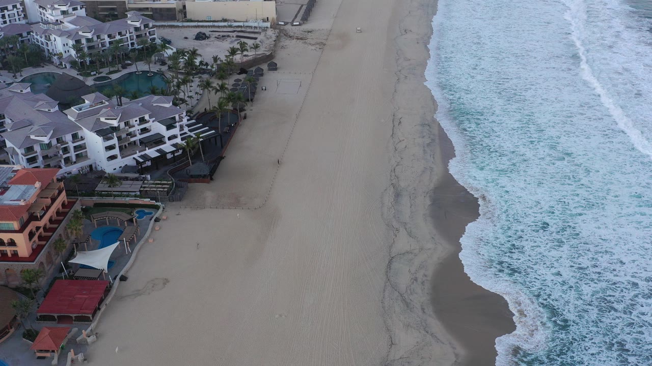 A high aerial frame over San Jose del Cabo shows resort buildings and pools at left, a wide empty sand strip in the center, foaming blue-green surf at right, and no visible people.