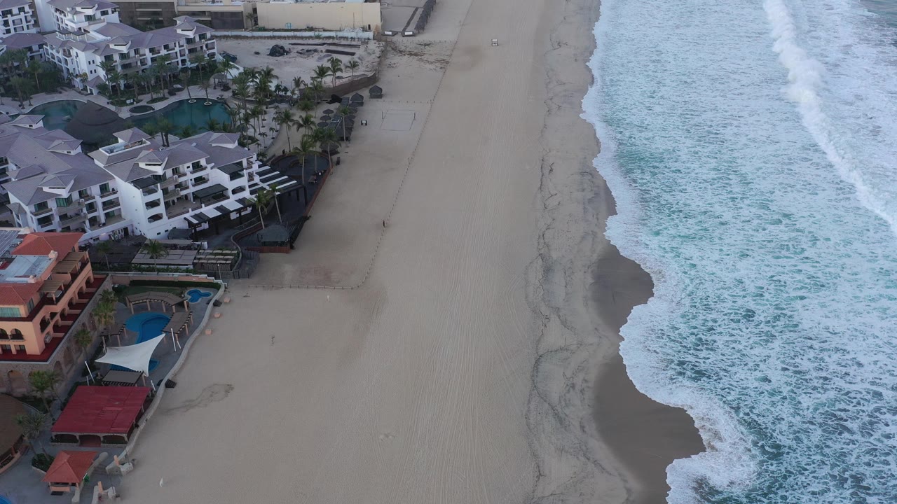 Overhead view of beachfront buildings, empty sand, and foamy ocean waves.