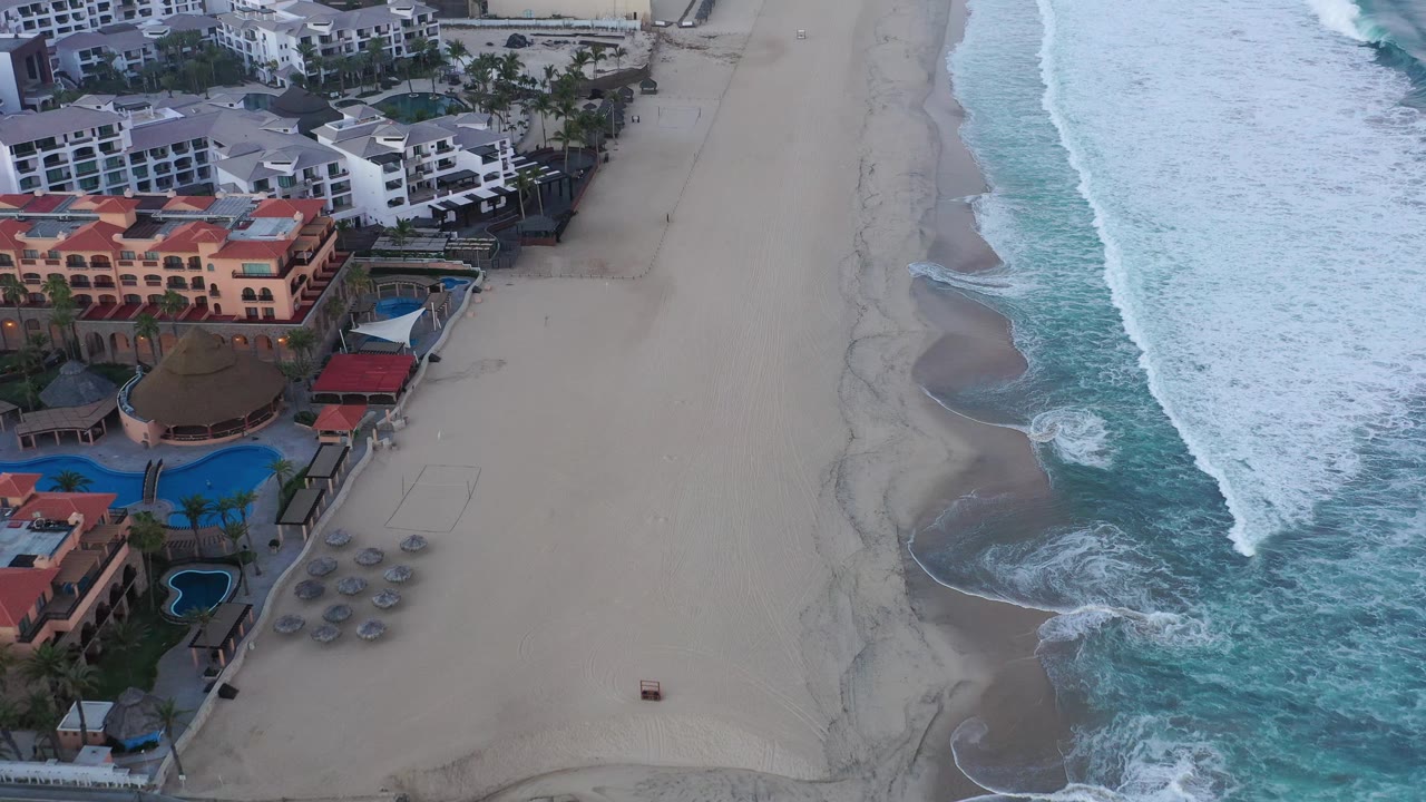 Aerial view in San Jose Del Cabo, Mexico with pale hotel buildings at the top, red-roofed resort structures at left, a wide sandy beach, curling surf, and no visible people.