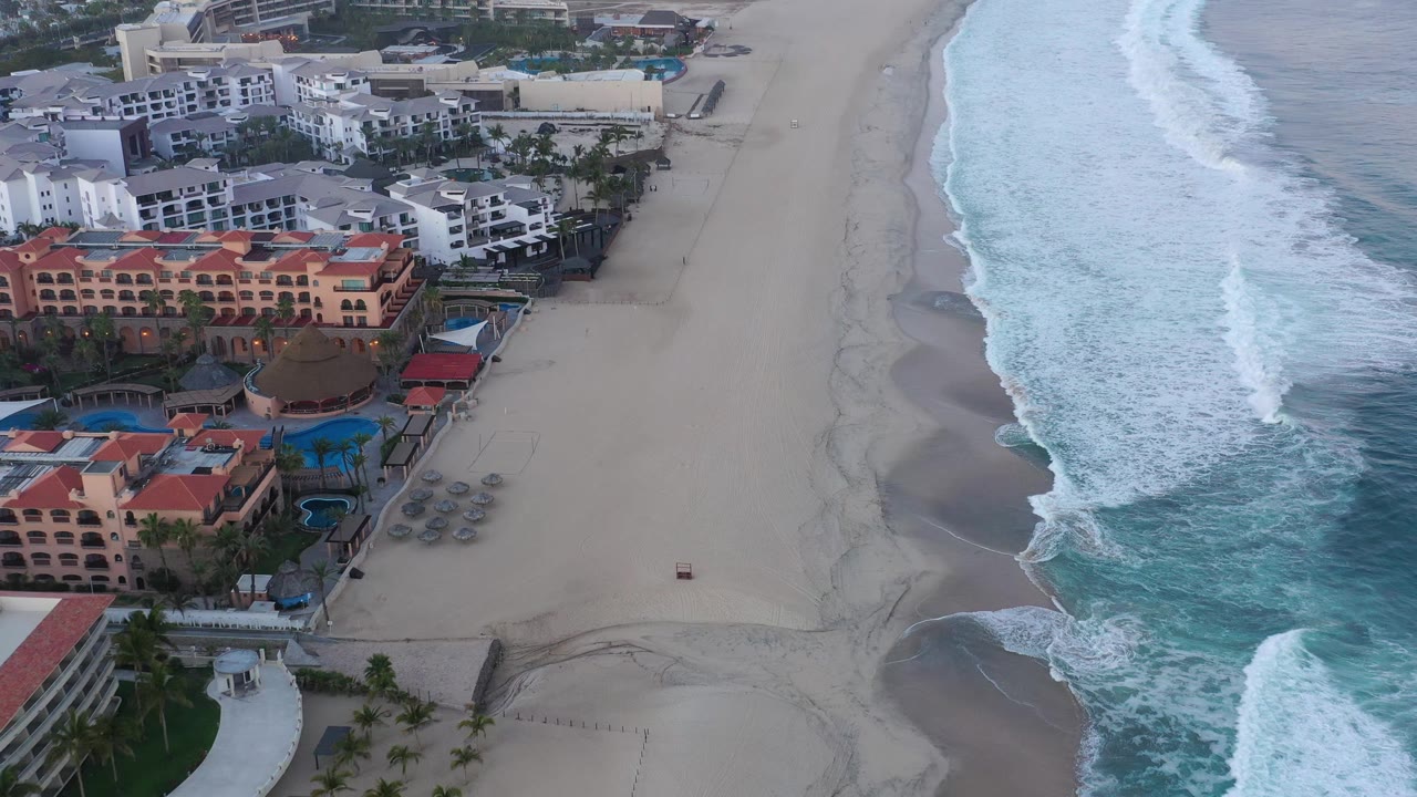 High aerial frame in San Jose del Cabo with the resort shifted across the left side, pale sand in the middle, and a long white wave band breaking near shore, with no visible people.