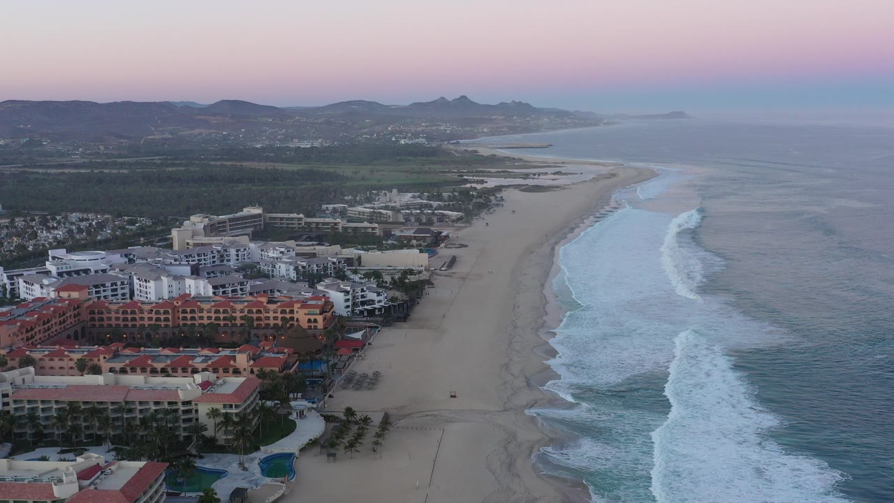 Aerial dusk view over San Jose del Cabo, Mexico, with red-roof resort buildings at lower left, a wide sandy beach, white surf lines, distant hills, and no visible people.