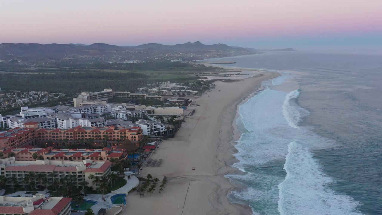 Aerial view in San Jose del Cabo, Mexico, looking along the beach as surf reaches the shore, hotels fill the lower left, hills sit in the background, and no visible people appear.