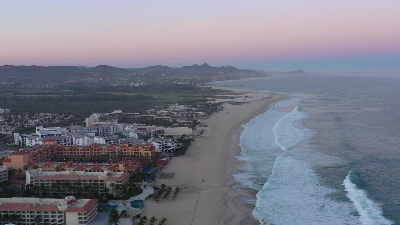 Aerial view of beachfront buildings, sandy coast, surf, and hills at dusk.
