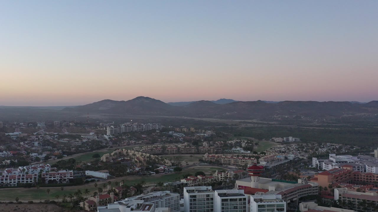 Opening aerial frame over San Jose del Cabo, Mexico, with foreground condos, a broad neighborhood grid, low hills on the horizon, pastel dusk sky, and no visible people.