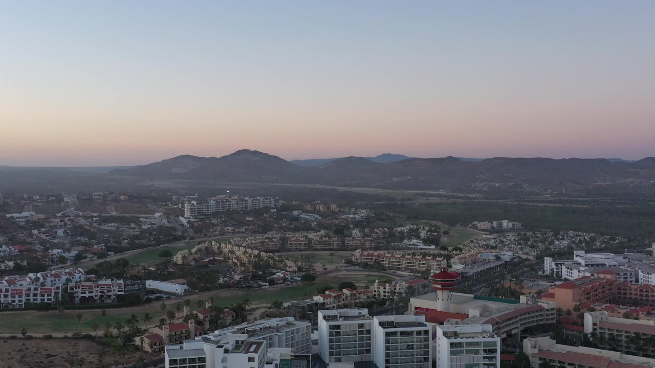 Dusk aerial with foreground rooftops, residential blocks, open land, and mountains.