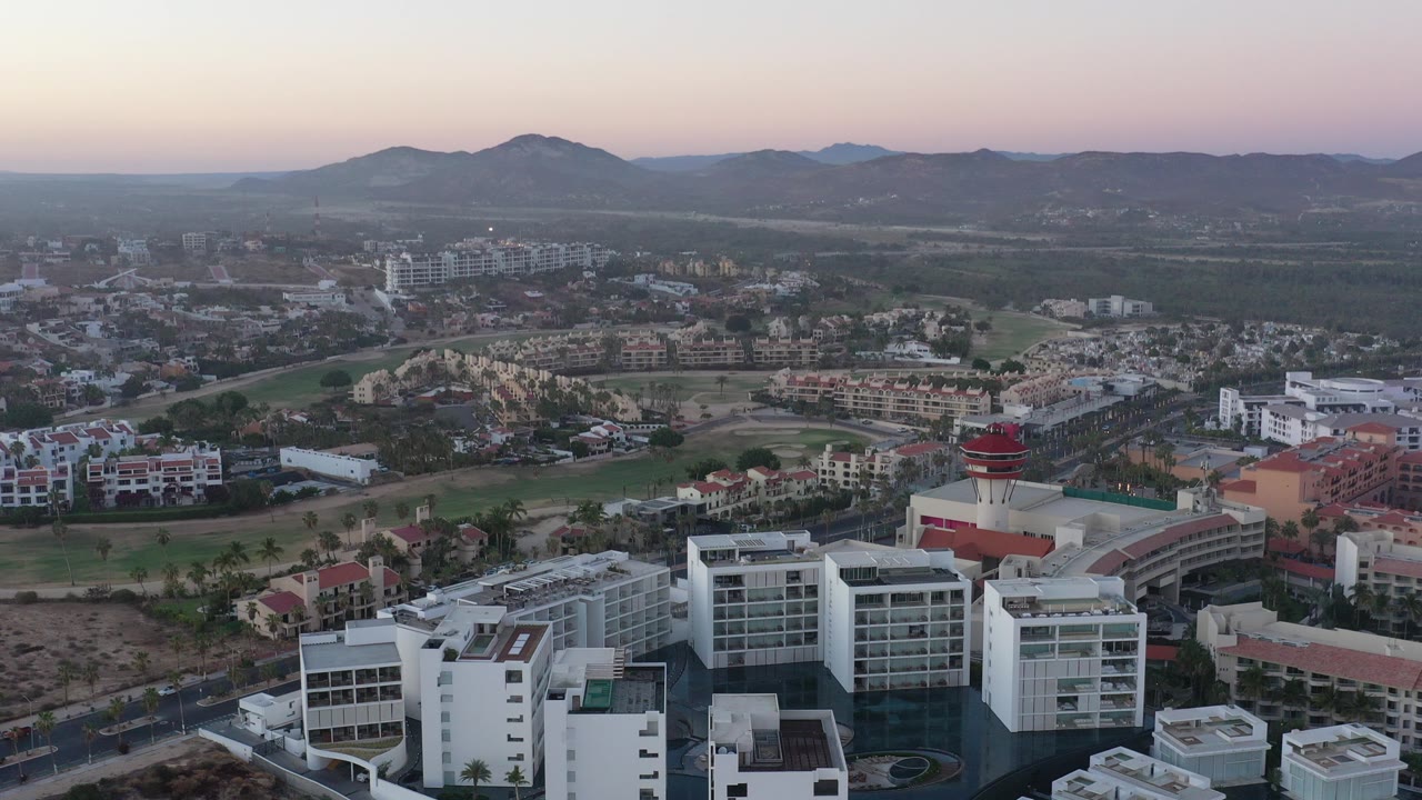 Opening aerial frame over San Jose del Cabo, Mexico, with white modern buildings in the foreground, township roads beyond, low mountains on the horizon, and no visible people.
