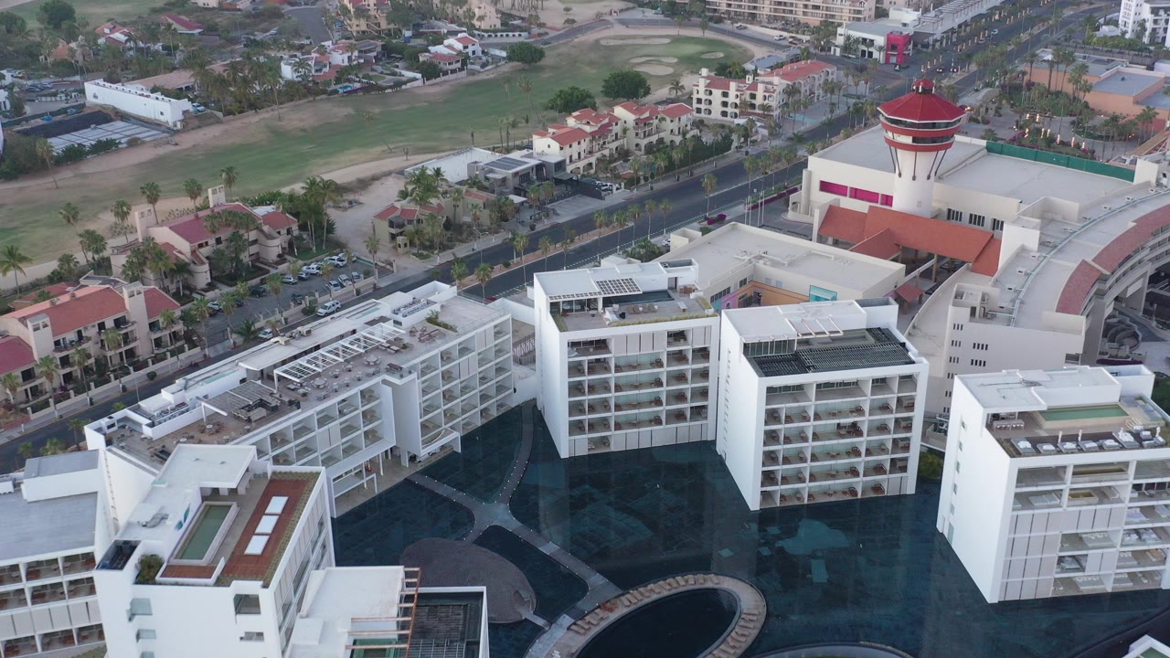 Aerial frame over San Jose del Cabo showing white multi-story buildings, a dark courtyard or pool area, nearby houses, a road, and no visible people.
