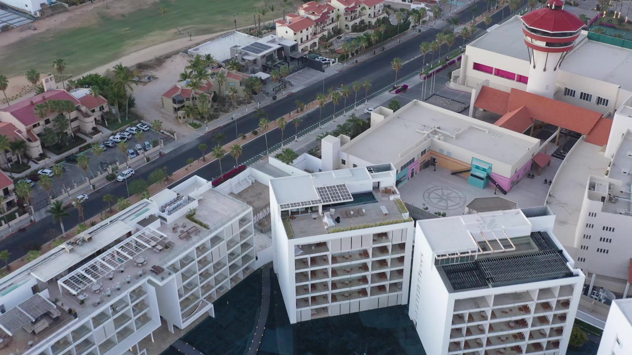 Aerial frame in San Jose del Cabo showing white hotel-like or apartment-style buildings, rooftop structures, a dark road lined with palms, parked vehicles, and no visible people.