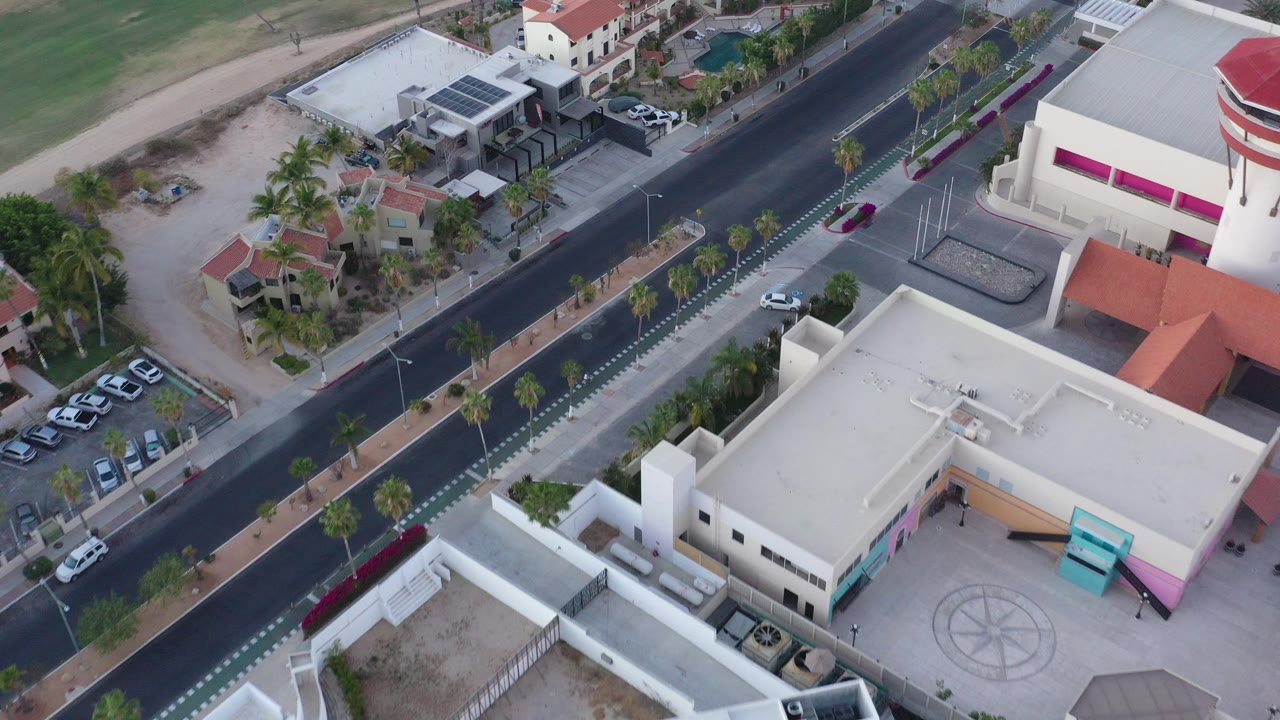 Oblique aerial view over San Jose del Cabo with a dark road, white rooftops, parked cars, palm trees, arid lots, and no visible people.