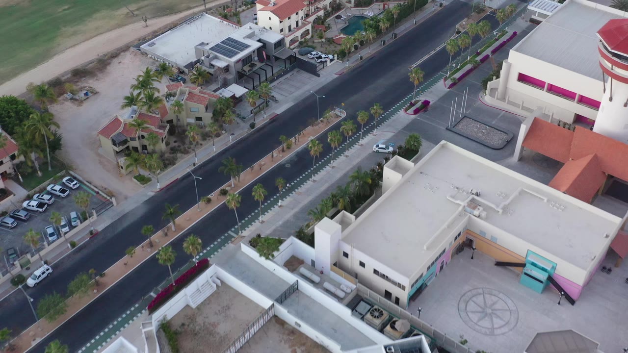 San Jose del Cabo aerial frame centered on the road and planted median, with white commercial-looking buildings, small residences, solar panels, and no visible people.