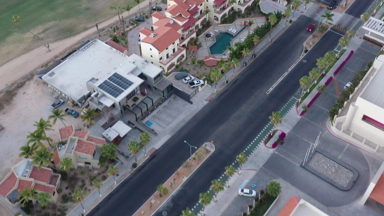 Aerial view of a paved road beside red-roof homes and a white building with solar panels.