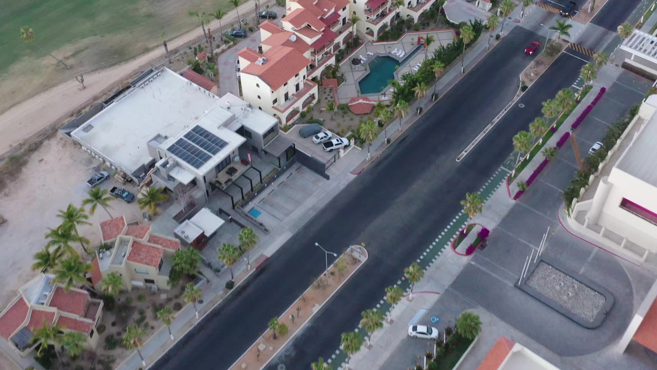 Aerial frame in San Jose del Cabo, Mexico, with the road centered between a white solar-panel building and red-roof residences, plus sidewalks, parked cars, palms, and no visible people.