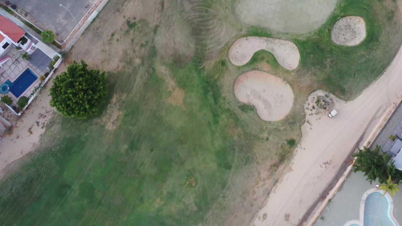 Overhead San Jose del Cabo frame showing fairway grass, multiple sand traps, a diagonal road edge, blue swimming pools, trees, and no visible people.