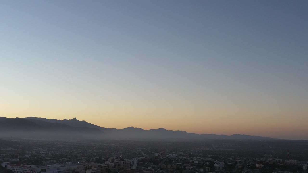 Wide aerial view over San Jose del Cabo with dark buildings below, a hazy mountain ridge on the horizon, pale evening sky above, and no visible people.
