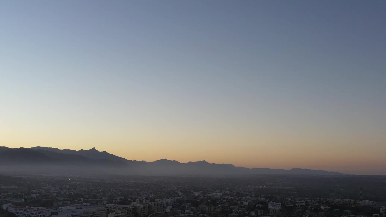 Aerial frame from San Jose del Cabo showing low town detail in shadow, layered mountains across the background, a warm dusk band near the horizon, and no visible people.