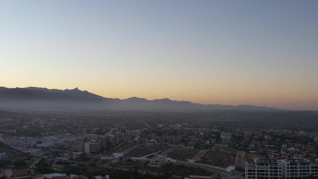 San Jose del Cabo appears from an elevated angle with scattered buildings, road grids, low haze, and a silhouetted mountain ridge, with no visible people.