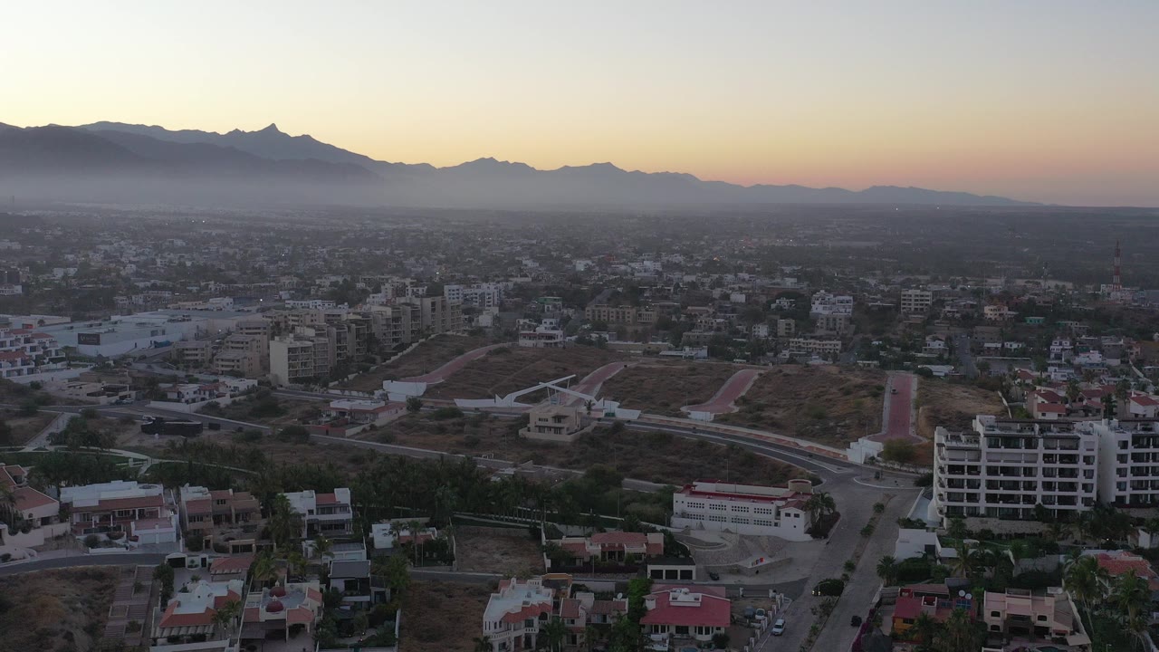 Aerial view over town buildings, dry hills, and distant mountains at sunset.