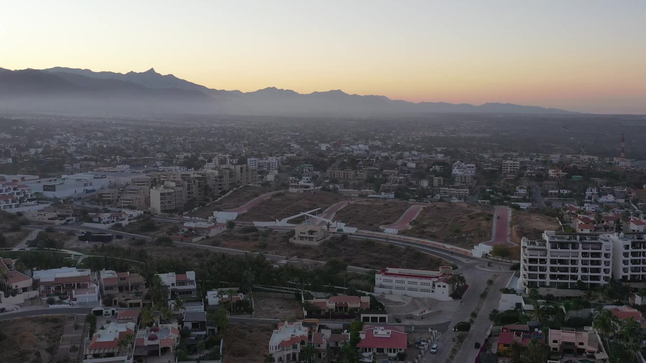 Second frame of San Jose del Cabo from above, showing a road junction, low-rise buildings, dry hillside lots, and the dark mountain ridge under warm dusk light, with no visible people.