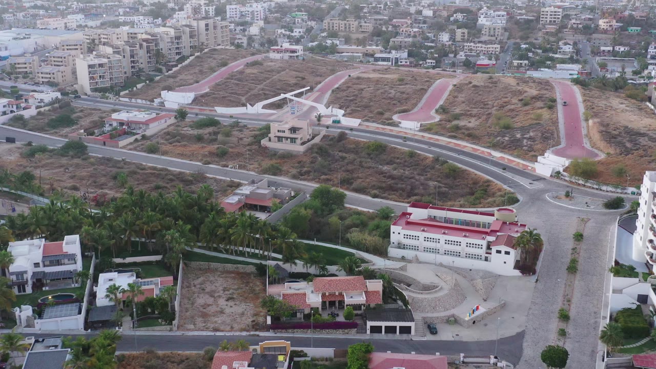 Oblique aerial frame in San Jose del Cabo showing a central road junction, palm grove, villas, dry hillside paths, distant buildings, and no visible people.