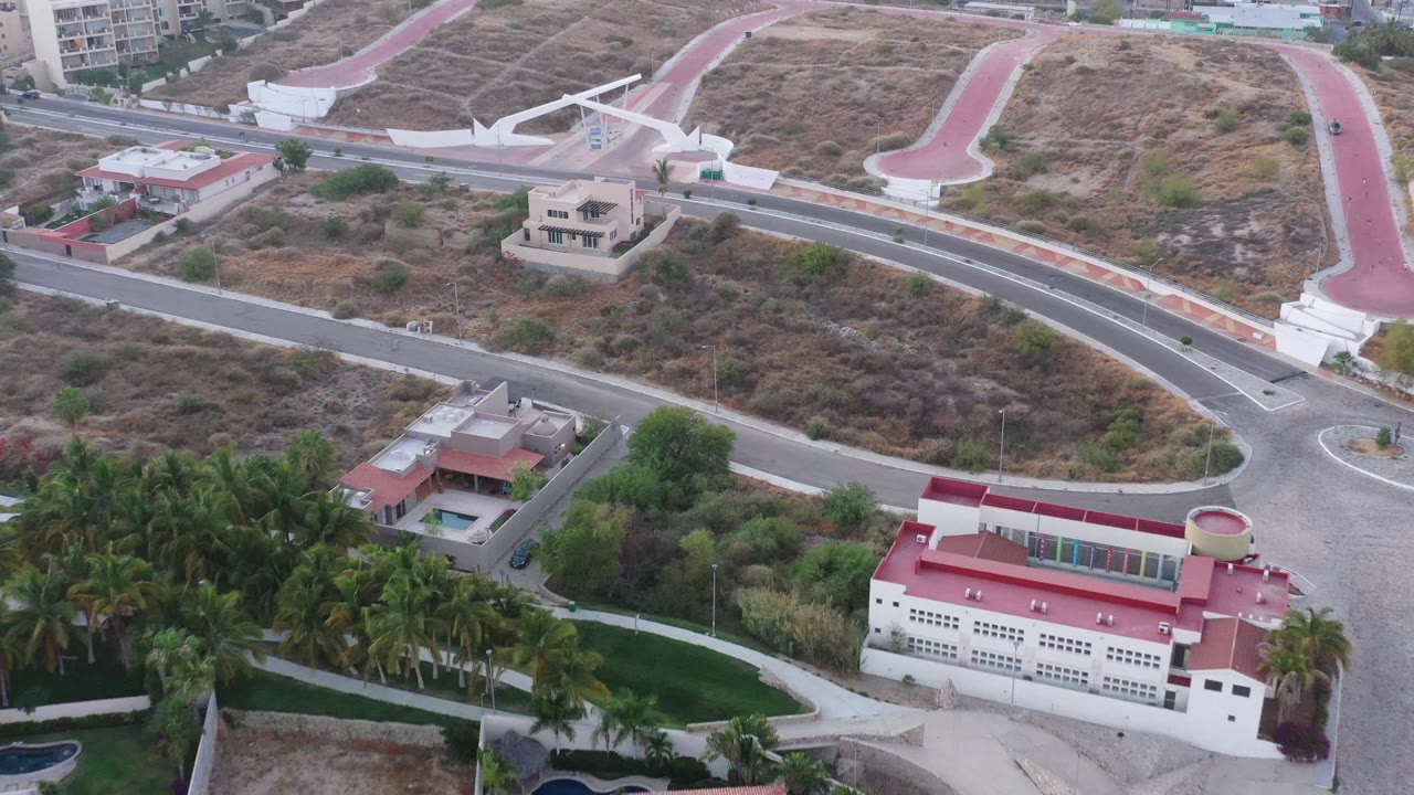 Aerial view in San Jose del Cabo showing a red-roofed building near the bottom edge, palms across the lower left, intersecting roads, dry brush, and no visible people.
