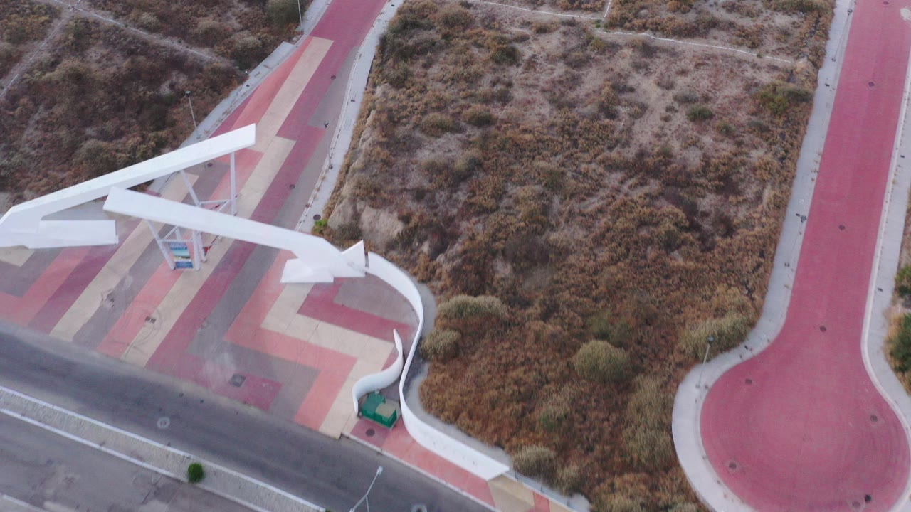 Top-down San Jose del Cabo frame showing a red paved path, tan geometric paving, dry scrubland, a white angular tower structure, and no visible people.