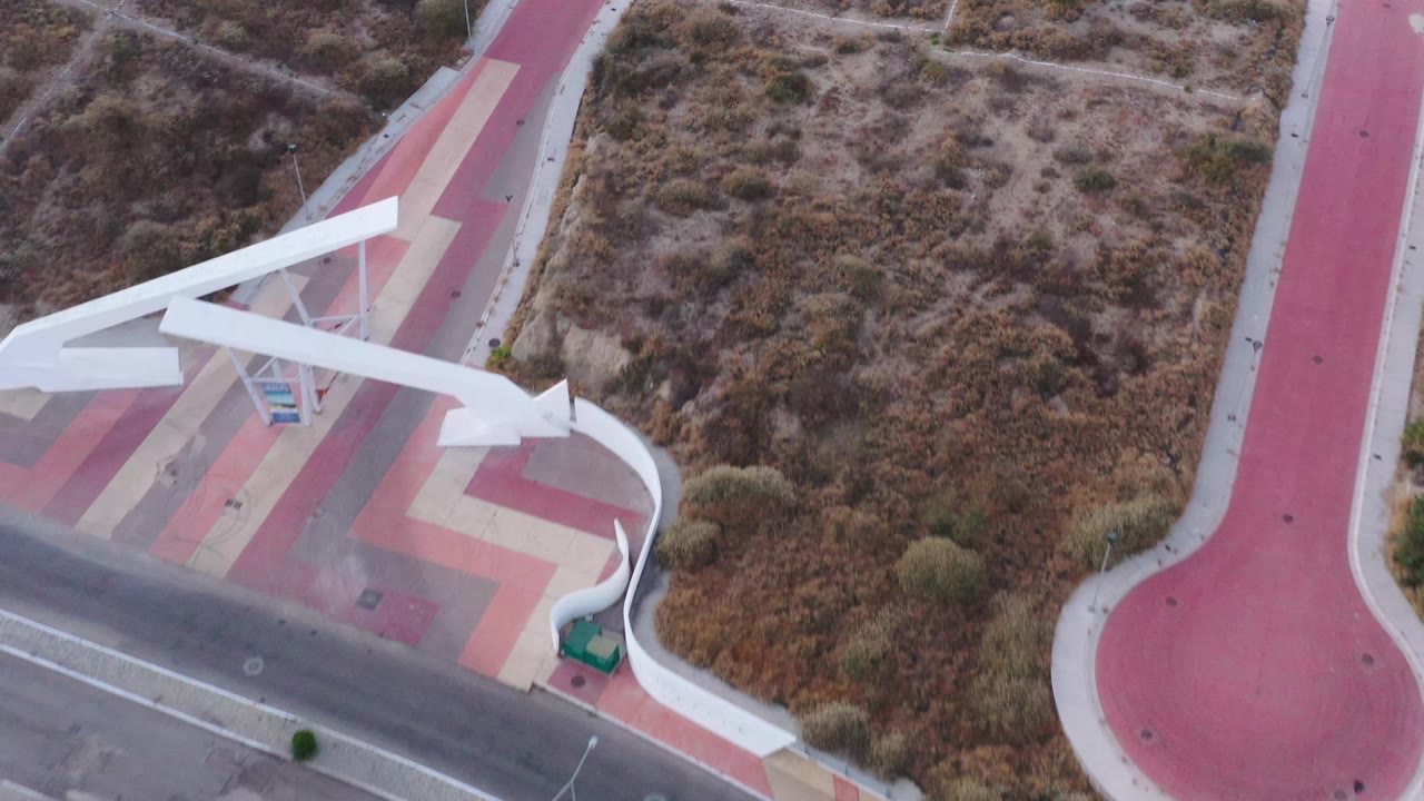 High overhead San Jose del Cabo view with the white tower crossing the lower-left frame, red path on the right, dry brush above, and no visible people.