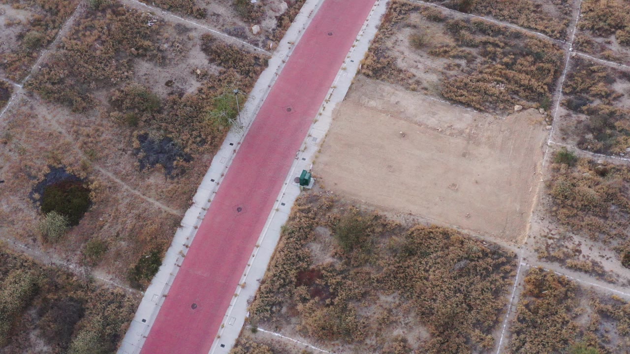 Top-down aerial frame in San Jose Del Cabo, Mexico, showing a red diagonal strip bordered by pale pavement, dry brush, dirt paths, and no visible people.