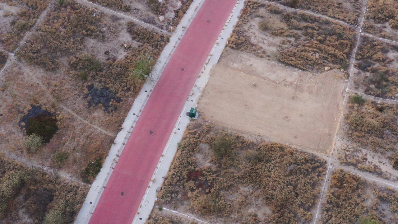 High aerial view in San Jose Del Cabo, Mexico, with the red strip on the left, a tan rectangular patch near center, scrub vegetation, crossing dirt paths, and no visible people.