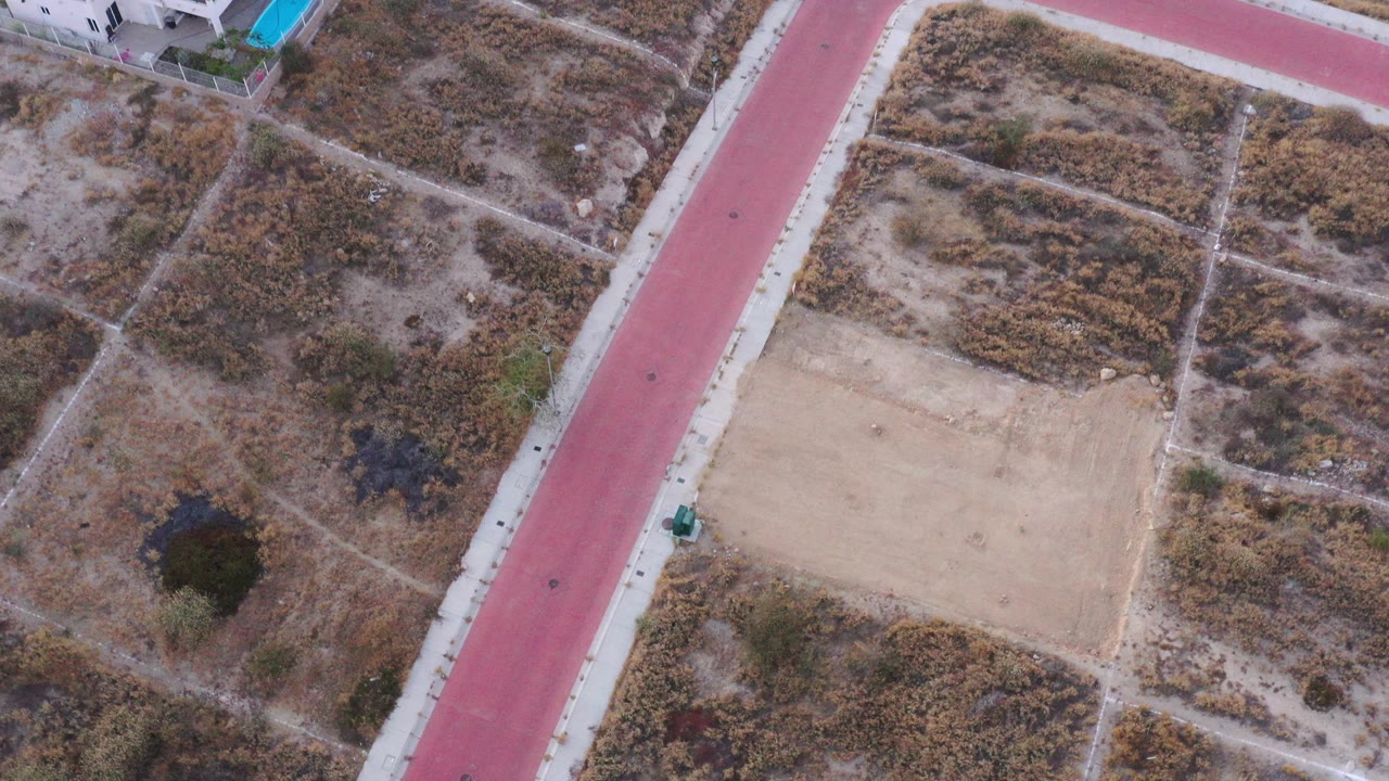 Overhead San Jose del Cabo frame showing a red paved road, pale sidewalks, dry scrub lots, one pale dirt rectangle, and no visible people.