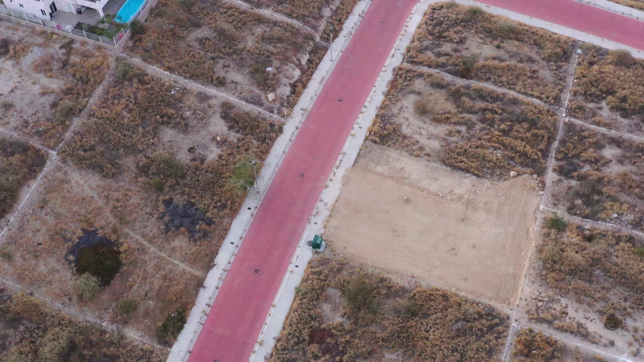 Aerial San Jose del Cabo view with the red road shifted left of center, scrub-covered parcels, a pale rectangular lot, scattered dark trees, and no visible people.