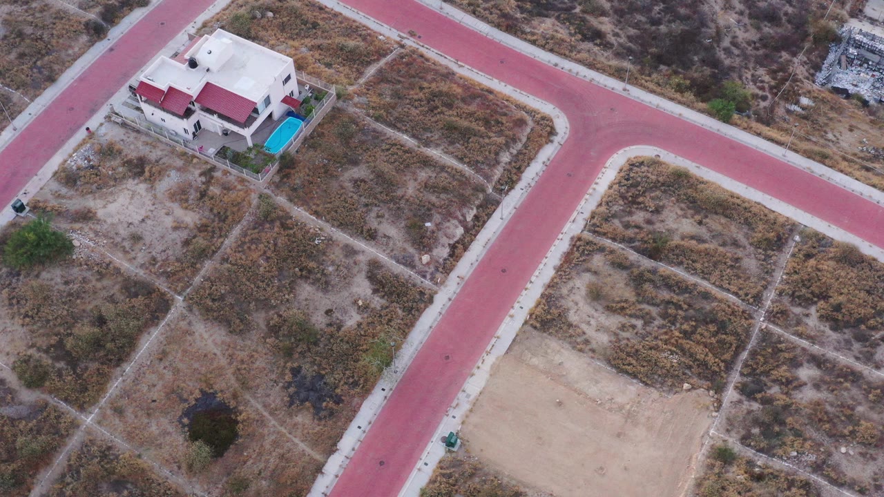 Aerial frame over San Jose del Cabo with the red road junction more centered, dry scrub parcels around it, the house and pool at upper left, and no visible people.