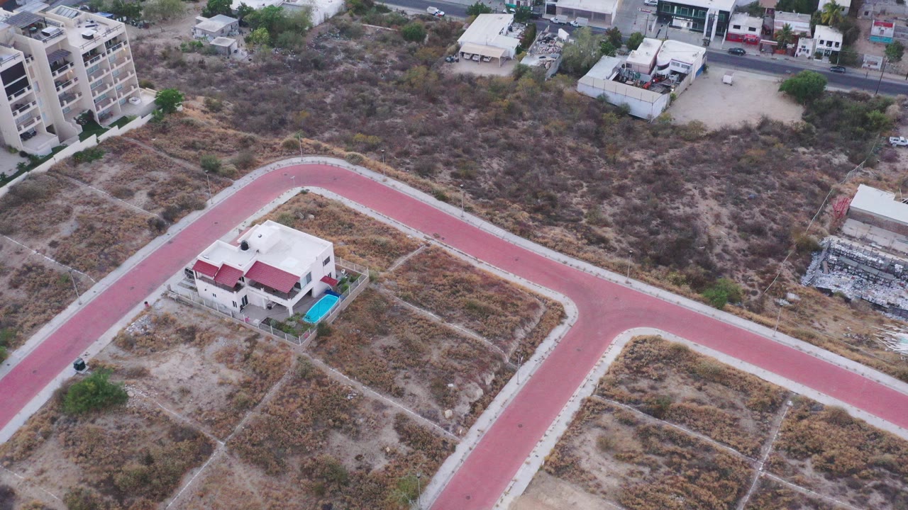 High aerial view in San Jose del Cabo showing a red paved road around scrubby empty plots, a small house with red roof sections, nearby apartments, and no visible people.