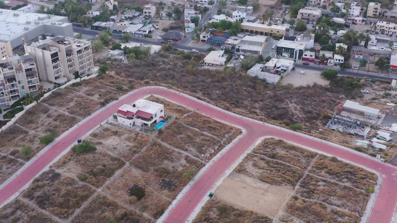 High aerial view over San Jose del Cabo showing a red curving road, dry scrub parcels, apartment buildings, and dense town blocks in the background, with no visible people.