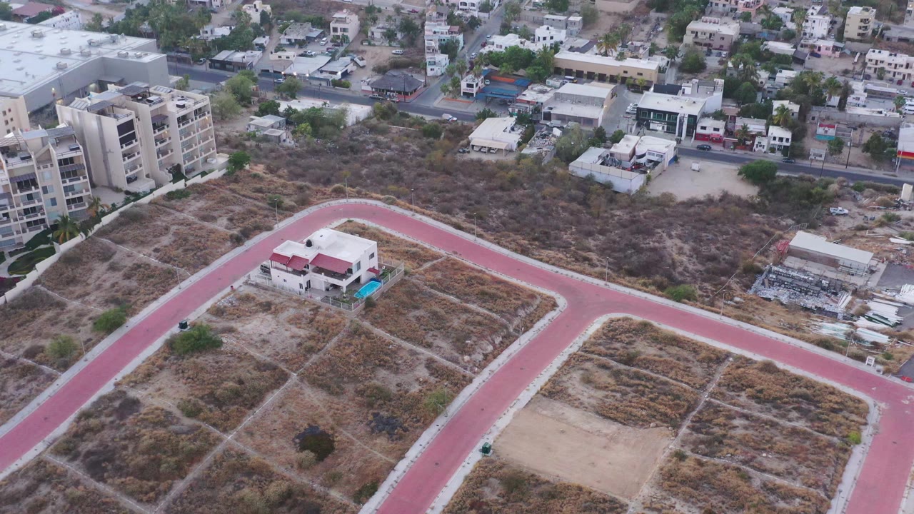 Aerial frame in San Jose del Cabo with the red road forming a broad bend around dry lots, a small red-roofed structure near center, and apartment blocks behind it, with no visible people.