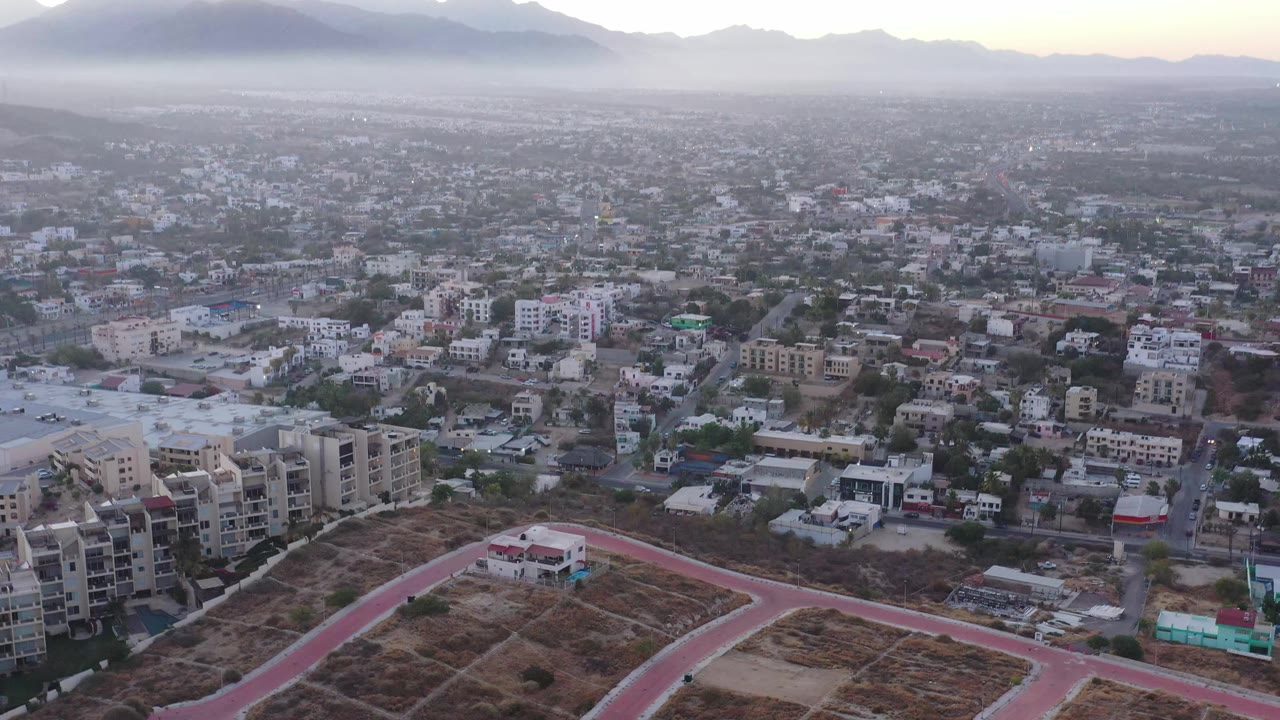 San Jose Del Cabo appears from above with apartment blocks, scattered homes, a dusty foreground lot, distant mountains, and no visible people.