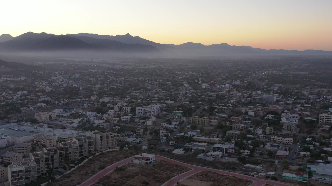 Aerial view over San Jose Del Cabo with dense city blocks below a hazy mountain ridge and warm sunset sky, no visible people.
