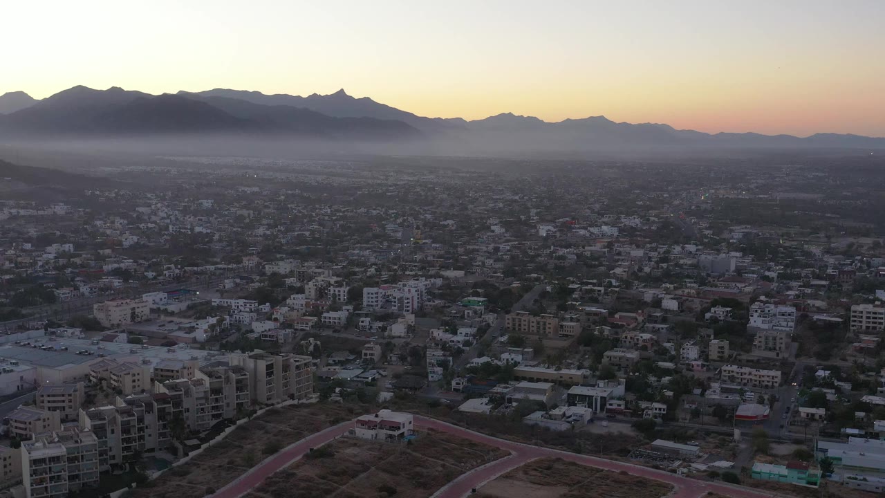 San Jose Del Cabo city rooftops, roads, and open lots sit beneath a broad mountain silhouette in fading evening light, no visible people.