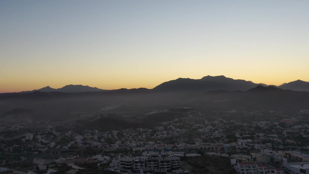 A wide San Jose del Cabo view shows clustered buildings low in frame, a hazy valley beyond them, layered mountain silhouettes, and no visible people.