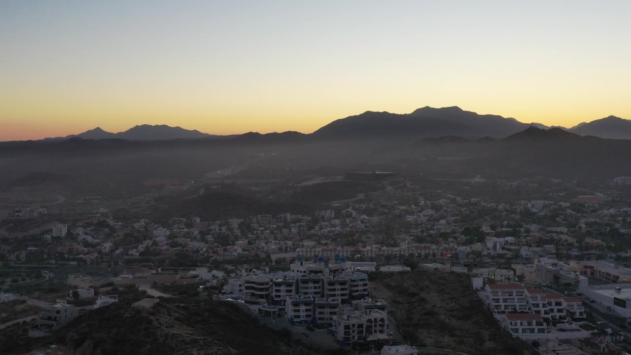 Opening frame of San Jose del Cabo from an elevated view, with hillside residences below, a hazy town valley, dark mountain silhouettes, warm sunset sky, and no visible people.