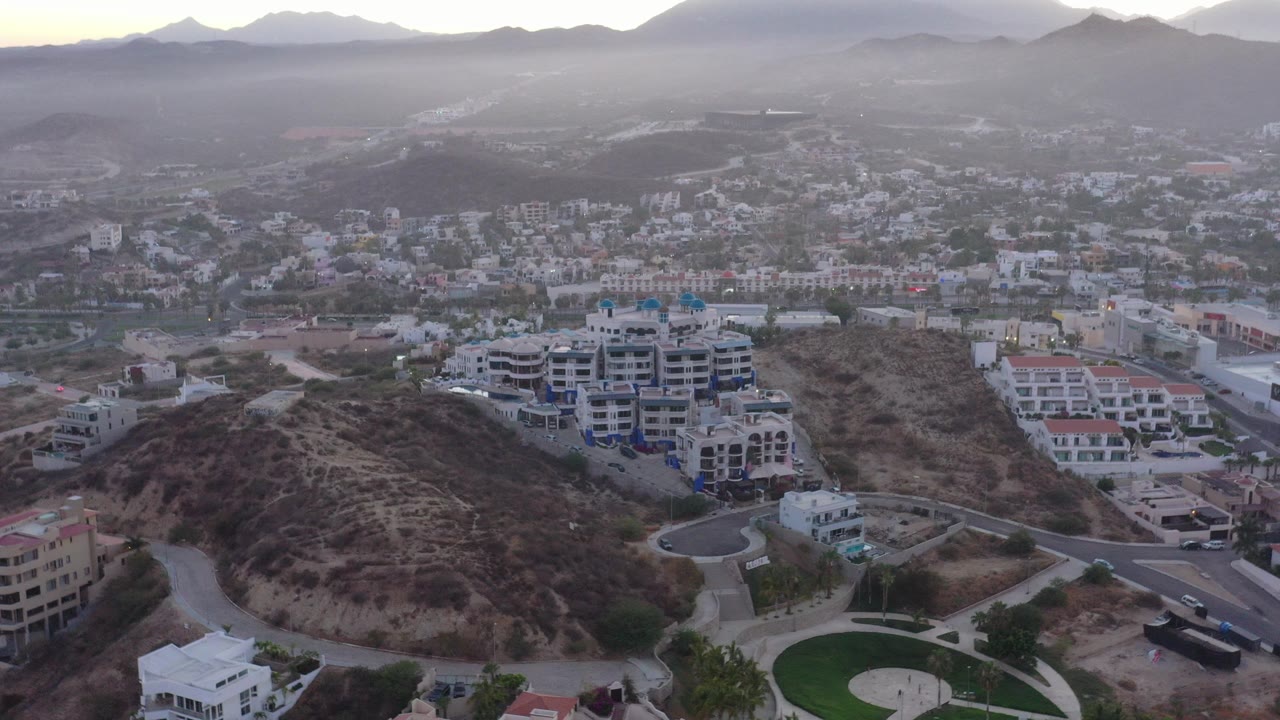 Aerial view over dry hills, roads, buildings, and distant mountains.