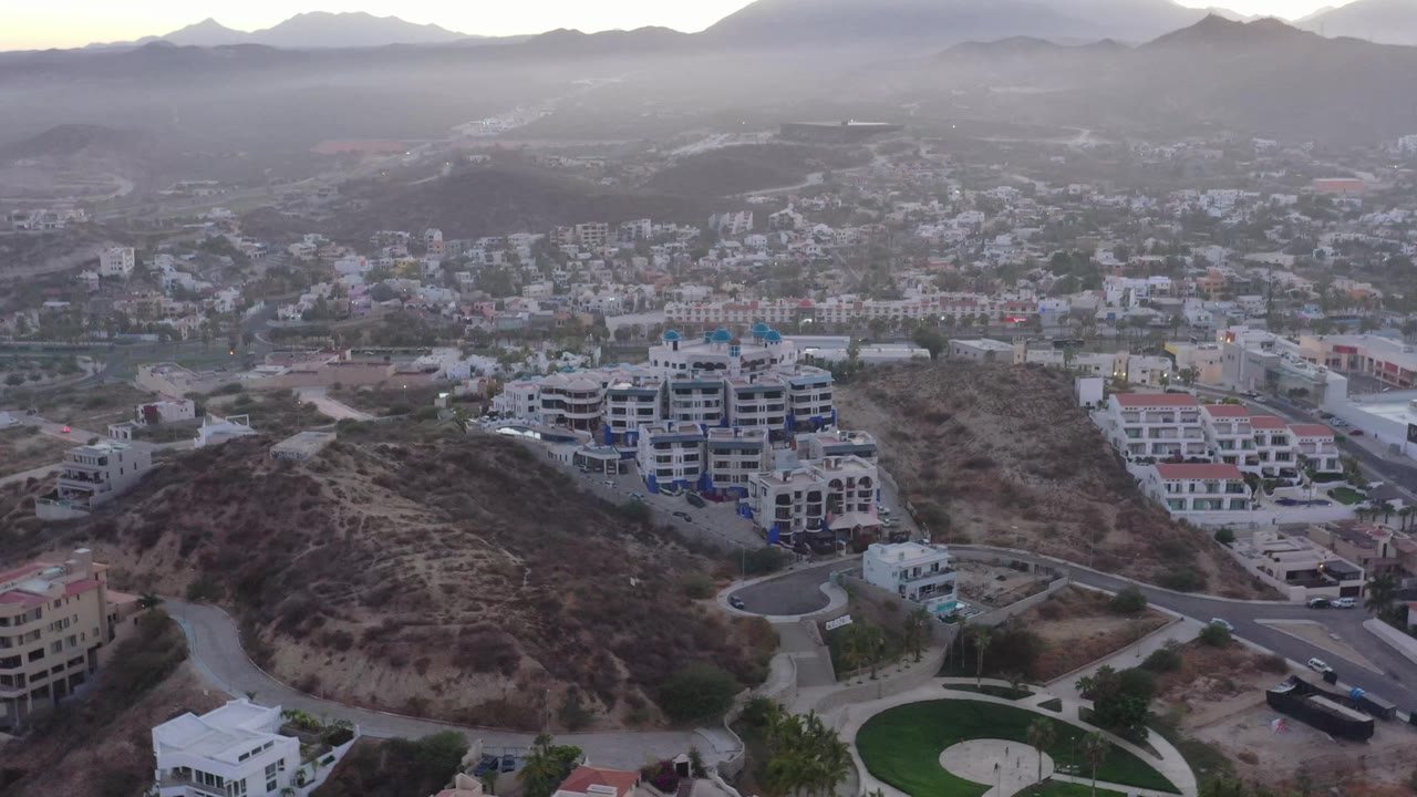 Aerial frame in San Jose del Cabo showing the scrub-covered hillside more centered, with white buildings, paved roads, a small circular green area, distant mountains, and no visible people.