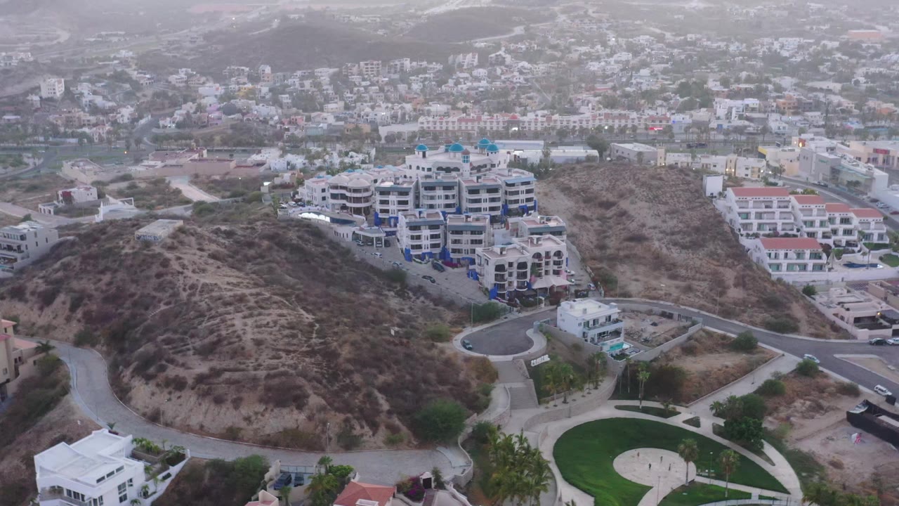 Aerial view in San Jose del Cabo, Mexico, showing curving roads around a brown hillside, modern white buildings, a small circular green landscaped area near the bottom edge, and no visible people.
