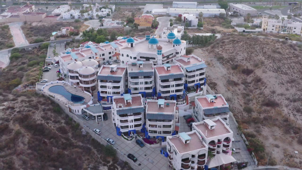 A slightly closer aerial angle shows the San Jose del Cabo complex with rounded balconies, blue-topped domes, a small pool area, paved drives, parked vehicles, and no visible people.