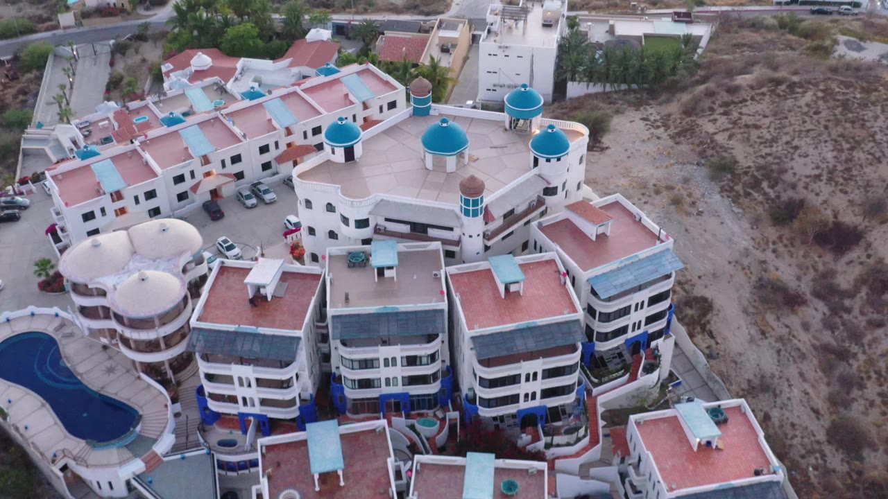 At 0:01, the San Jose del Cabo aerial angle remains over the complex, centering the red roofs, white facades, blue rooftop domes, parked cars, and no visible people.