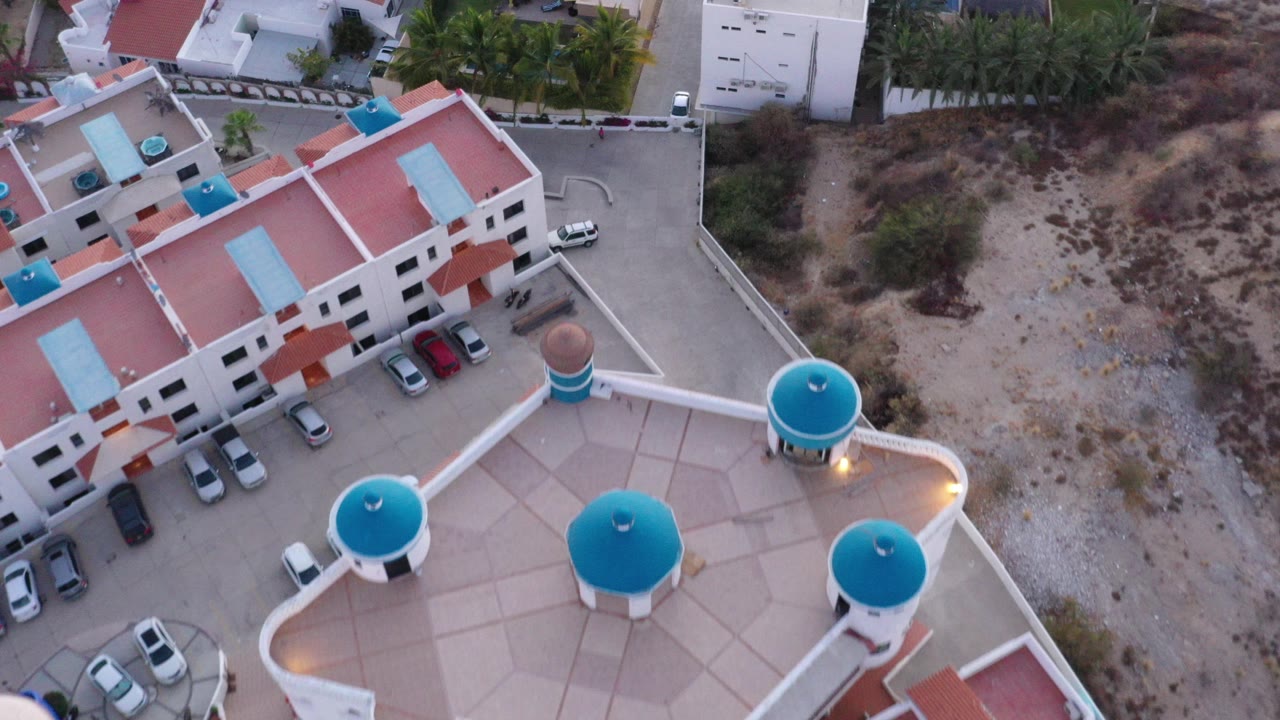 High drone view over San Jose del Cabo residential buildings with turquoise rooftop domes, red tile roofs, parked cars, dry brush beyond the road, and no visible people.