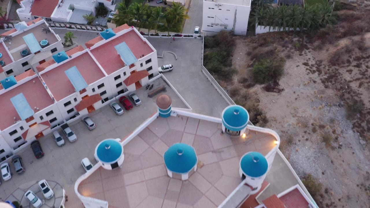 Aerial frame in San Jose del Cabo showing the residential complex shifted across the lower left, with parked cars along the building, palm trees near the road, dry open ground, and no visible people.