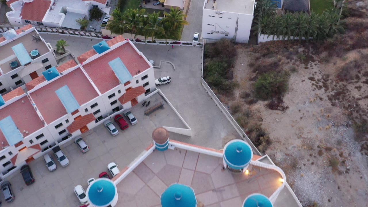 Overhead residential scene with red roofs, turquoise rooftop features, parked cars, road, palms, and brush.