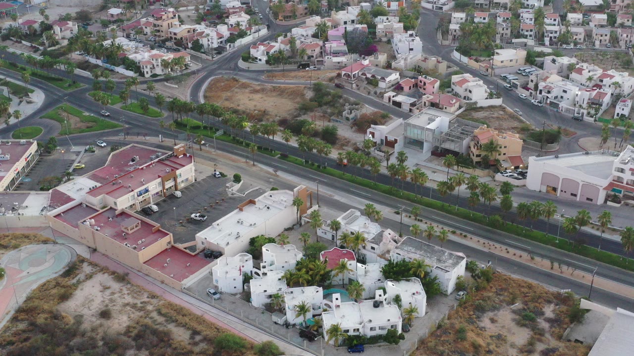 High aerial view over San Jose del Cabo with white homes in the foreground, a divided road across the center, commercial buildings, residential blocks, dry lots, and no visible people.